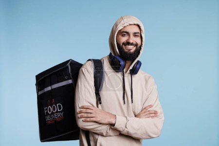 Téléchargez les photos : Homme arabe avec une expression joyeuse portant un sac à dos et offrant un portrait de la nourriture. Jeune livreur de restaurant souriant portant capuche debout avec sac thermique et regardant la caméra - en image libre de droit