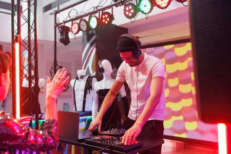Téléchargez les photos : Musicien souriant mélangeant pistes électroniques avec station de DJ pour les personnes qui font la fête dans une boîte de nuit. Jeune homme se produisant sur scène alors que les clubbers s'amusent et dansent en club avec des projecteurs - en image libre de droit