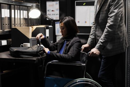 Foto de Gerente ejecutiva asiática empresaria con discapacidad en silla de ruedas y compañera de trabajo empezando turno de trabajo. Contadores rodeados de papeleo de datos burocráticos en la oficina del gabinete de depósito - Imagen libre de derechos