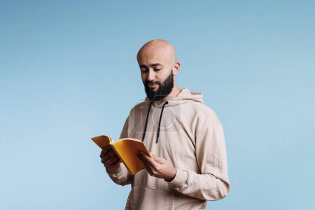 Photo for Arab man holding open book and reading novel with carefree expression. Young smiling bald bearded arabian person in hoodie standing with yellow softcover notepad and learning - Royalty Free Image