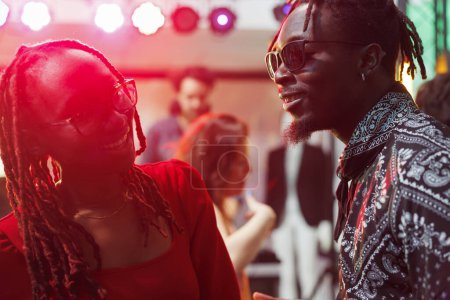 Téléchargez les photos : Couple afro-américain souriant riant et bavardant dans une boîte de nuit sombre éclairée de lumières vibrantes. Homme et femme insouciants parler et socialiser tout en faisant la fête dans le club - en image libre de droit