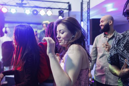 Foto de Mujer joven festejando y bailando en un club nocturno lleno de gente mientras asistía a un evento de reunión social. Clubber chica moviéndose a ritmo de música en vivo en la pista de baile rodeado de gente - Imagen libre de derechos