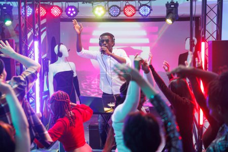 Téléchargez les photos : Le DJ afro-américain se produit sur scène et parle à la foule dansante au micro. Les gens se détendent et se déplacent sur la piste de danse à la discothèque de musique électronique partie dans la boîte de nuit - en image libre de droit
