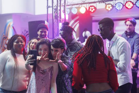 Téléchargez les photos : Copines souriantes prenant selfie sur smartphone tout en faisant la fête avec des amis dans une boîte de nuit. Femmes gaies posant pour la photo de téléphone portable tout en s'amusant à la discothèque dans le club - en image libre de droit