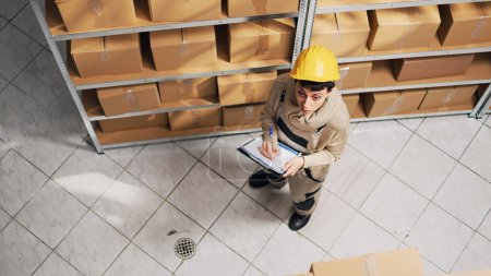 Photo for Female supervisor checking list of products on racks, looking at cardboard boxes to plan distribution. Woman with hardhat working in warehouse with merchandise and stock, storage inventory. - Royalty Free Image