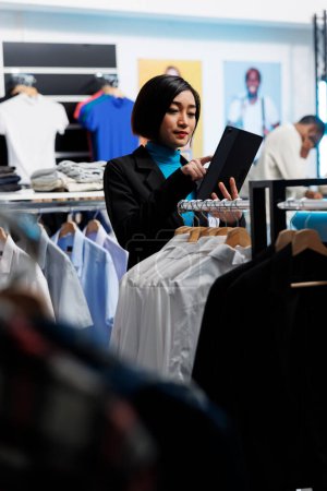 Téléchargez les photos : Magasin de vêtements asiatique femme employée utilisant tablette numérique tout en travaillant dans le centre commercial. Ouvrier du centre commercial du département examinant les vêtements en stock et gérant les stocks - en image libre de droit