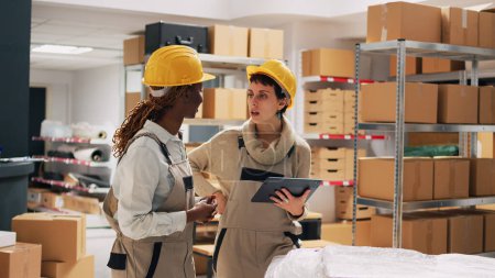 Photo for Two women with hardhats organizing products on shelves, looking at tablet to check stock logistics and planning distribution. Warehouse manager and employee counting cardboard packs. - Royalty Free Image