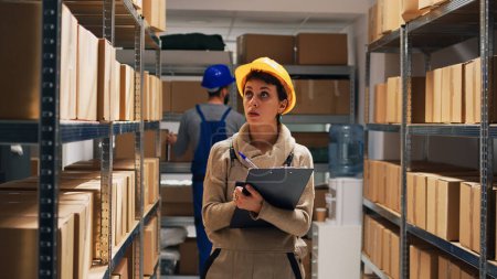 Photo for Depot supervisor checking list of merchandise on files, using papers to do cargo inventory. Woman in overalls and hardhat counting number of boxes and packages on warehouse racks. - Royalty Free Image