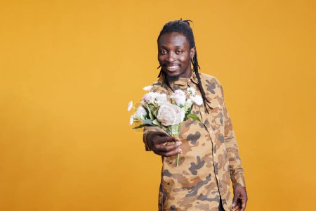 Téléchargez les photos : Portrait de l'homme afro-américain tenant bouquet de fleurs blanches, présentant surprise pour la petite amie à la caméra en studio sur fond jaune. copain romantique posant pour la Saint-Valentin - en image libre de droit