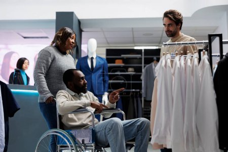 Foto de Hombre afroamericano con discapacidad crónica que recibe consejos de un asistente de tienda de ropa mientras hace compras con un amigo. Cliente del centro comercial del departamento en silla de ruedas hablando con un consultor - Imagen libre de derechos