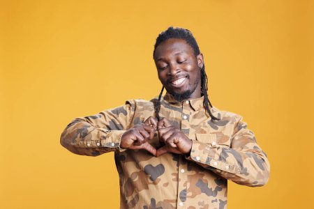 Photo for Smiling adult doing heart shape with hands on camera, promoting love in front of camera in studio over yellow background. African american excited lovely person expressing love feelings and gratitude - Royalty Free Image