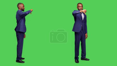 Photo for Negative office worker doing thumbs down in studio, wearing formal suit over greenscreen background. Young businessman showing dislike gesture and expressing disapproval on camera. - Royalty Free Image