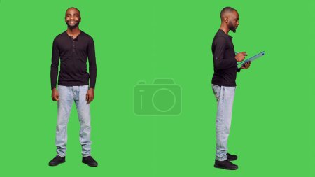 Photo for Confident man posing on camera and taking notes, writing important information using pen and clipboard papers. Casual male model standing over greenscreen bacground, leisure activity. - Royalty Free Image