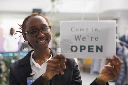 Foto de Tienda de ropa sonriente mujer afroamericana asistente colgando letrero de apertura en la puerta de entrada. Alegre trabajador de la tienda de moda poniendo letrero en la ventana notando el comienzo del día de trabajo - Imagen libre de derechos