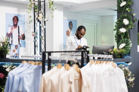 Photo for Clothing store african american employee working at counter, planning sales and writing in organiser. Shopping mall fashion department assistant standing at checkout desk - Royalty Free Image
