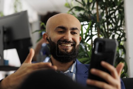 Téléchargez les photos : Gestionnaire exécutif arabe souriant téléconférence à l'aide d'un smartphone tout en travaillant dans un bureau d'affaires. Startup entrepreneur faire la gestion de l'équipe virtuelle sur vidéocall de téléphone mobile - en image libre de droit