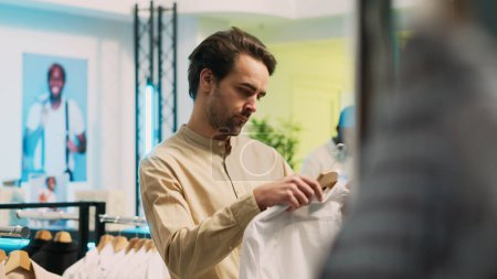 Photo for Caucasian man looking at fashionable wear on hangers, buying modern clothes on sale in shopping center. Male customer examining fashion boutique merchandise, commercial activity. Handheld shot. - Royalty Free Image