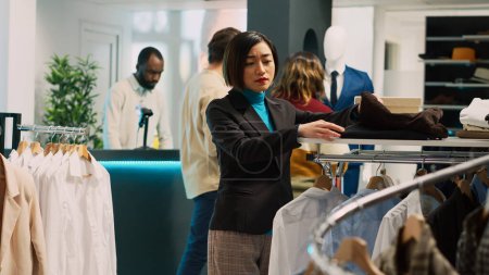 Photo for Asian woman shopping in fashionable clothing store, looking at formal and casual wear on hangers. Female customer buying trendy merchandise from shopping center, retail shop. Tripod shot. - Royalty Free Image