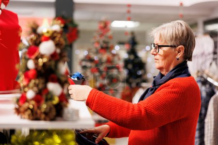 Photo for Senior woman browsing for accessories and fashion items in festive retail store, looking for gifts and christmas dinner outfit. Customer checking material of clothing on racks at shopping mall. - Royalty Free Image