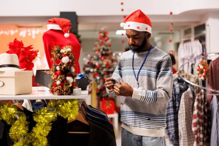 Photo for Store assistant wearing santa hat arranging clothes on racks for seasonal deals and promotions, Christmas decorated clothing store. Supervisor looking at accessories in retail shop during xmas. - Royalty Free Image