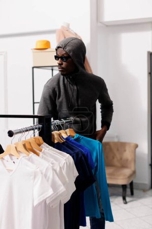 Foto de Ladrón afroamericano eligiendo robar mercancía de moda, mirando a su alrededor para ver si alguien lo observa. Ladrón robando boutique moderna, usando capucha y gafas de sol. Concepto de crimen - Imagen libre de derechos