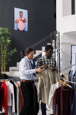 Foto de Cliente afroamericano revisando la camisa de moda, discutiendo el material de ropa con el empleado en la boutique moderna. Hombre adicto a las compras para trajes de descuento, la compra de ropa casual. Concepto de moda - Imagen libre de derechos