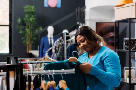 Foto de Mujer afroamericana que comprueba la tela de la camisa en boutique moderna, comprando mercancías de moda. Cliente Shopaholic compra ropa de desgaste formal en la tienda de ropa. Concepto de moda - Imagen libre de derechos