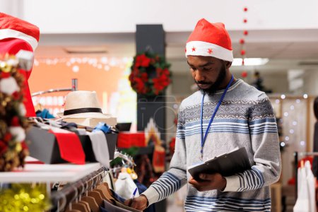 Photo for African american man counting hangers in clothing store, working on stock inventory while he wears santa hat. Retail sales assistant checking list of merchandise in shopping center mall. - Royalty Free Image