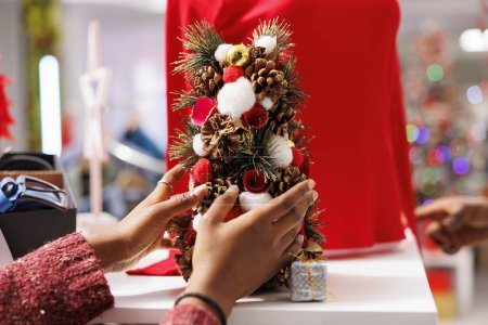 Photo for Woman employee arranging small tree festive decor all around clothing store near racks and hangers, personnel decorating christmas ornaments. African american girl putting decorations up. Close up. - Royalty Free Image