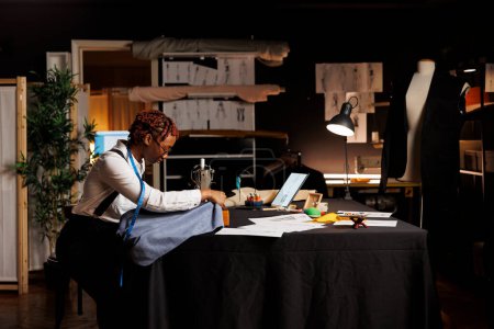 Téléchargez les photos : Couturière afro-américaine utilisant une machine à coudre industrielle pour faire de la broderie créative sur un morceau de tissu à utiliser sur la prochaine collection de mode sur mesure. Expert tailleur travaillant sur la collection sartoriale - en image libre de droit