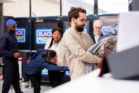 Photo for Young man shopper holding clothes in hands while standing at clothing store checkout, people paying for Black Friday purchases. Male customer enjoying discount shopping during sales - Royalty Free Image