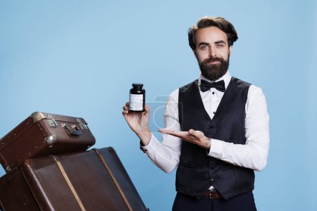 Photo for Smiling doorman presents bottle of pills in studio, doing marketing ad for medicaments and supplements. Professional bellboy holding jar filled with vitamins or drugs next to luggage. - Royalty Free Image