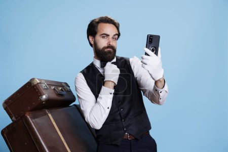 Photo for Elegant hotel employee takes photos while being on studio camera, posing with confidence and being dressed in formal hotel attire. Professional stylish bellhop using smartphone for pictures. - Royalty Free Image