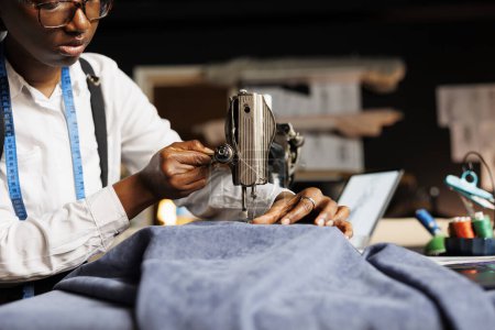 Foto de Primer plano de la costurera afroamericana utilizando la máquina de coser industrial en material de tela en el estudio de sastrería. Suitmaker corte prendas de vestir pieza, creando atuendo a medida - Imagen libre de derechos