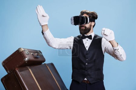 Photo for Male bellhop enjoying vr headset interactive vision, taking care of lugagge and posing on camera. Professional doorkeeper in formal suit using virtual reality concept, hotel staff. - Royalty Free Image