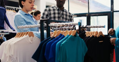 Photo for Stylish employee showing black shirt to couple, checking new fashion collection in modern boutique. African american customers shopping for trendy merchandise and accessories in clothing store - Royalty Free Image