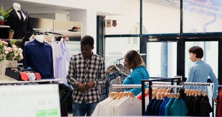 Photo for African american couple shopping for formal wear, checking trendy clothes fabric before buying it in modern boutique. Stylish people looking at hangers with new fashion collection in clothing store - Royalty Free Image