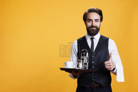 Photo for Happy waiter does thumbs up sign while he holds platter with coffee and accessories in studio, professional stylish butler. Man restaurant employee showing like agreement gesture on camera. - Royalty Free Image