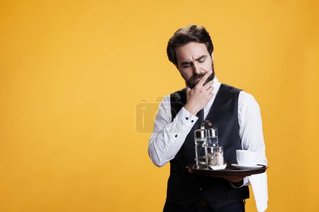 Photo for Luxury butler thinking about ideas, preparing to serve customers with food or drinks in yellow studio. Stylish person with waiter occupation being thoughtful with tray in hand on camera. - Royalty Free Image