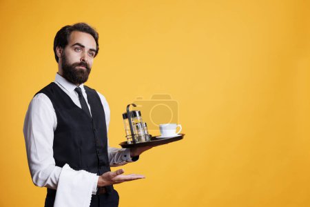 Photo for Skilled elegant butler pointing aside and holding food platter to serve customers, working at five star restaurant. Young adult dressed in suit and tie carries tray with cup of coffee. - Royalty Free Image