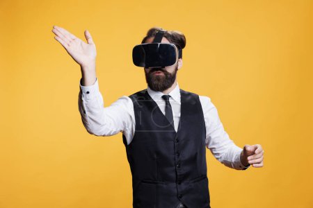Photo for Young butler using virtual reality headset, enjoying 3d simulation on modern headset against yellow background in studio. Catering employee having fun with interactive vision 3d glasses. - Royalty Free Image