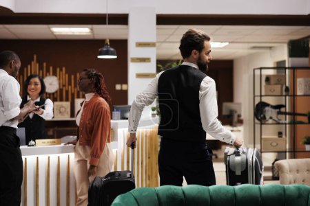 Photo for Hotel bellboy helping guests with bags, carrying luggage to room in luxury resort. Young man working as concierge giving assistance to people with suitcases arriving at reception. - Royalty Free Image