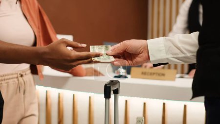 Photo for Close up of thankful hotel guest tipping helpful bellboy after receiving great customer service. African american woman handing dollar banknote to porter employee for helping with baggage - Royalty Free Image