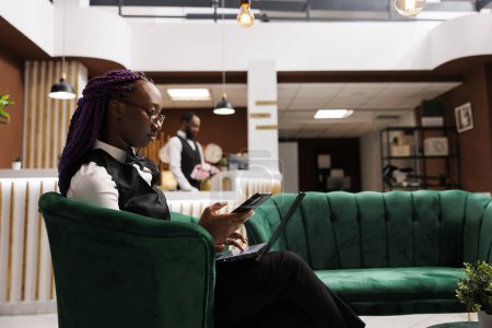 Téléchargez les photos : Jeune femme afro-américaine gestionnaire d'hôtel assis à la réception salon en utilisant smartphone et ordinateur portable, la gestion du budget et la commande de fournitures. Technologie dans l'industrie hôtelière - en image libre de droit