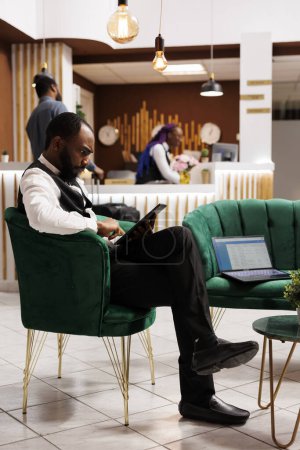 Téléchargez les photos : Focalisé gestionnaire d'hôtel concentré tenant tablette numérique à l'aide du logiciel de réception, confirmant les réservations en ligne tout en étant assis dans le hall. Technologies innovantes dans l'industrie hôtelière - en image libre de droit