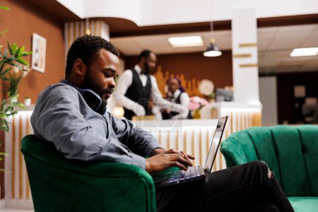 Téléchargez les photos : Focalisé homme afro-américain pigiste dactylographier sur clavier d'ordinateur portable en utilisant le wifi de l'hôtel, assis dans le hall, travailler à distance ou étudier en ligne tout en voyageant. Télétravail et voyages d'affaires - en image libre de droit