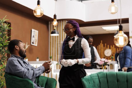 Photo for Smiling friendly African American woman hotel employee in uniform talking and interacting with happy guest in lobby. Generous rich man traveler giving cash tip to waitress girl at resort - Royalty Free Image