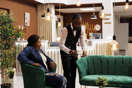 Téléchargez les photos : Serveur d'hôtel de type africain servant du café aromatique chaud à une femme assise dans le hall. Femme noire voyageur commandant cappuccino en attendant l'enregistrement à la réception, Caféine et décalage horaire - en image libre de droit