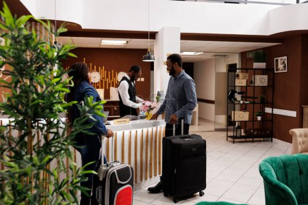 Photo for Happy black couple standing at hotel front desk with luggage, checking in at honeymoon resort, people waiting at reception for check-in. Receptionist helping guests with check out - Royalty Free Image