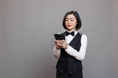 Téléchargez les photos : Serveuse de restaurant asiatique souriante en noir et blanc message de dactylographie uniforme sur smartphone. Service traiteur attrayant femme employée utilisant un téléphone mobile tout en posant dans le studio - en image libre de droit
